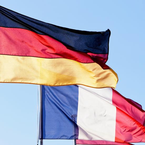 Et tysk og et fransk flag blafrer i vinden mod blå himmel.