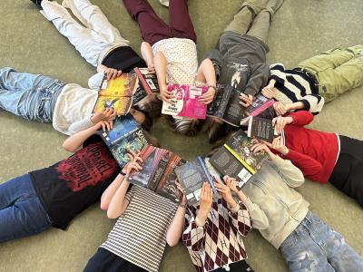 Readathon. Unge ligger i en cirkel med hovederne mod hinanden og ansigterne dækkede af åbne bøger.