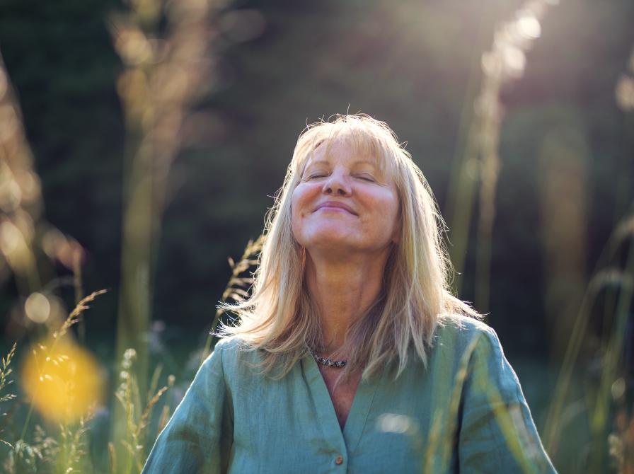 En kvinde står med lukkede øjne og ser ud til at nyde øjeblikket på en græseng omgivet af træer en forårs- eller sommerdag badet i sollys.