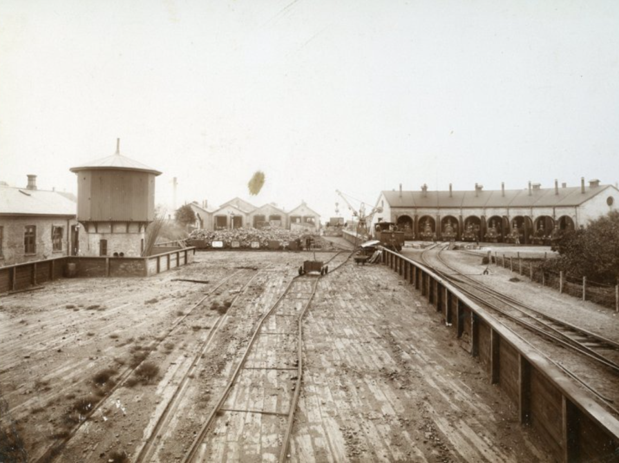 Rundremise med plads til 8 lokomotiver med tilhørende drejeskive ses til højre i billedet. I baggrunden midt i billedet ses værkstedsbygningerne, hvoraf en del stadig er bevaret og taget i brug af Nyborg Destilleri. Tårnet til venstre i billedet er et vandtårn, hvorfra damplokomotiverne påfyldtes det nødvendige vand til dampproduktion. 1890.